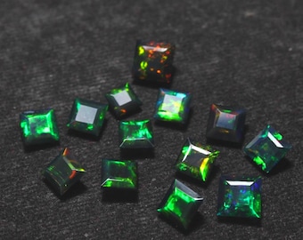Puede incluir: Una colección de ópalos negros de corte cuadrado con vibrantes destellos verdes, azules y naranjas. Las piedras preciosas están dispersas sobre una superficie gris oscuro, mostrando sus cualidades iridiscentes. Adecuado para la creación de joyas.