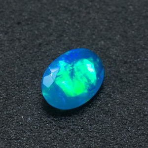 Puede incluir: Ópalo ovalado con un vibrante juego de colores. La gema presenta una base azul brillante con destellos verdes y una superficie facetada. El ópalo se encuentra sobre un fondo oscuro y texturizado.