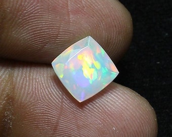 Pode incluir: Uma gema de opala branca com um corte quadrado de almofada. A opala apresenta um arco-íris de cores, incluindo verde, amarelo e azul.