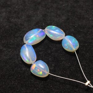 May include: Strung oval-shaped opals with iridescent flashes of color. The opals are a milky white with internal play-of-color, strung on a thin white cord. The gemstones are smooth and polished, showcasing their natural beauty.