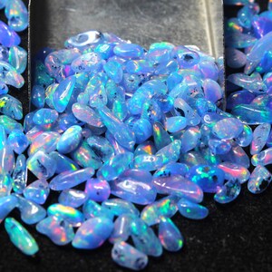 May include: A close-up of a collection of polished opals. The opals are a light blue colour with iridescent flashes of pink, green, and purple. The gemstones are displayed in a metal scoop against a dark background.