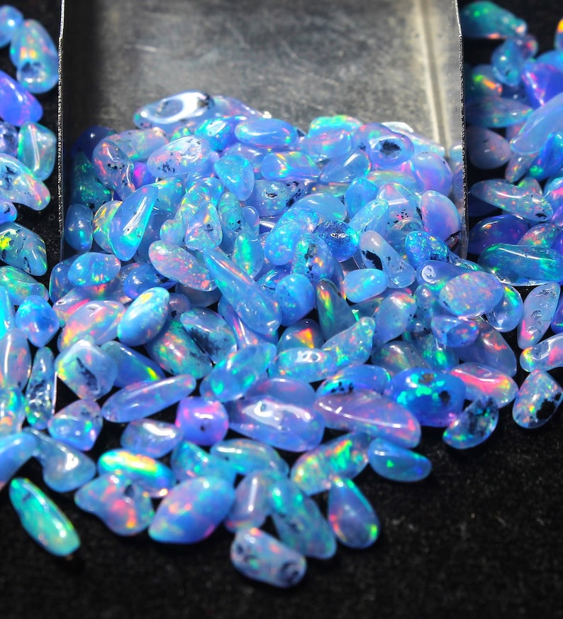 May include: A close-up of a collection of polished opals. The opals are a light blue colour with iridescent flashes of pink, green, and purple. The gemstones are displayed in a metal scoop against a dark background.