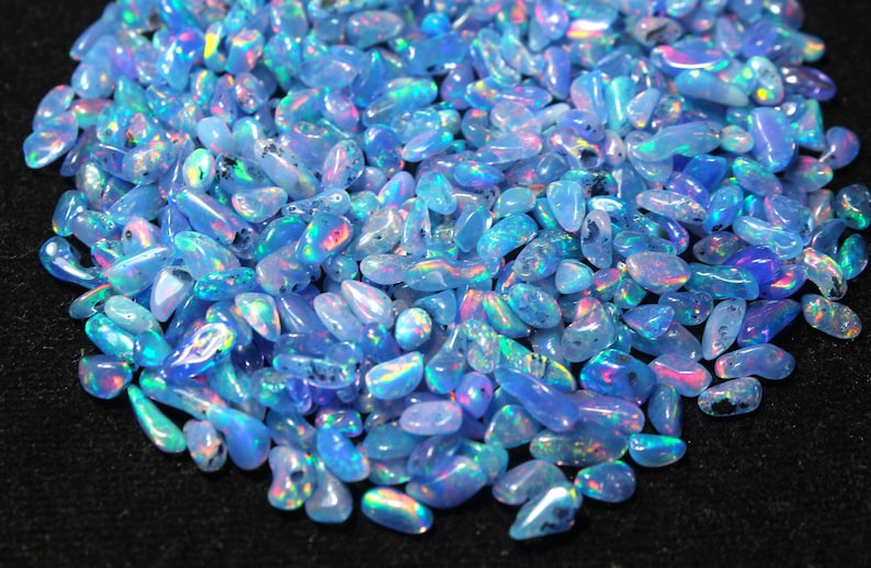 May include: A collection of polished opals, predominantly light blue with iridescent colours. The gemstones are oval-shaped and vary in size. The opals have a smooth, glossy surface with flashes of pink, green, and yellow. The background is black.