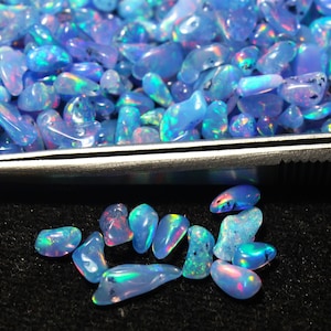 May include: A selection of polished, iridescent blue opal gemstones. The opals are in various shapes and sizes, with a mix of light and dark blue hues. Some stones display flashes of colour, including pink, green, and yellow. The opals are on a black surface.