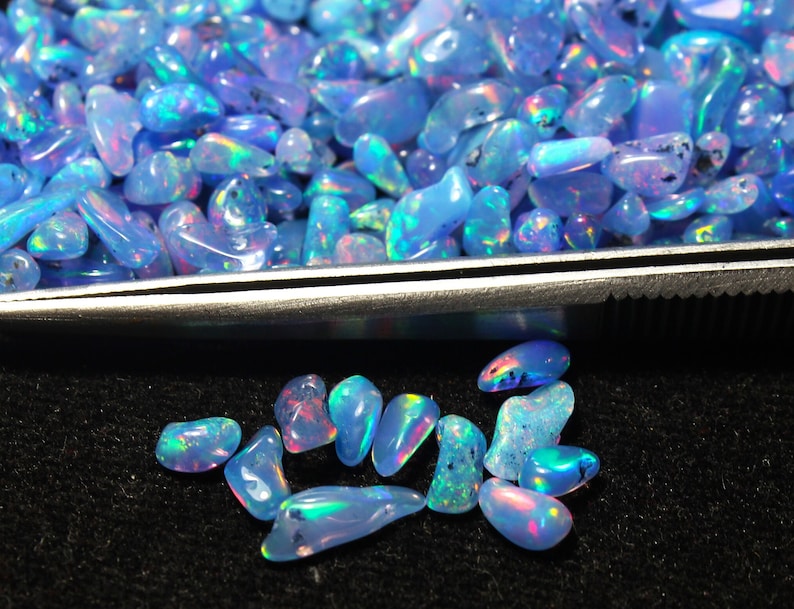 May include: A selection of polished, iridescent blue opal gemstones. The opals are in various shapes and sizes, with a mix of light and dark blue hues. Some stones display flashes of colour, including pink, green, and yellow. The opals are on a black surface.