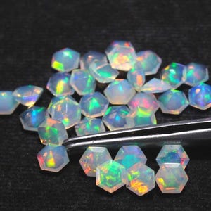 May include: A collection of iridescent, hexagonal-cut opal gemstones. The opals display a play of color with flashes of green, blue, and orange. A pair of tweezers holds one of the opals, suggesting they are loose stones.