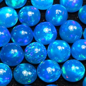 May include: A collection of round, blue opal cabochons with iridescent flashes of color. The opals are smooth and polished, and they are arranged in a random pattern.