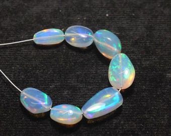 May include: A strand of oval-shaped opal beads. The opals are a milky white color with iridescent flashes of blue, green, and yellow. The beads are strung on a thin, white string, and are displayed against a black background.