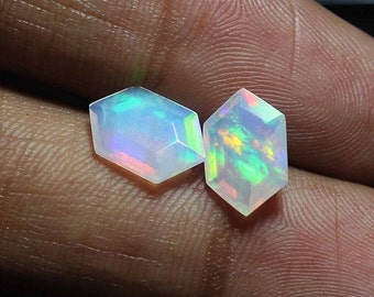 May include: Two iridescent opal gemstones, cut into a hexagonal shape with faceted edges. The opals are a pale white with flashes of green, blue, and yellow.