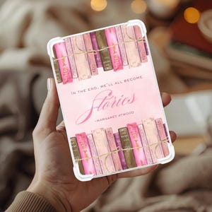 May include: A white tablet case featuring a pink design with stacked books and the text "In the end, we'll all become Stories - Margaret Atwood." The design includes pink, brown, and purple books tied with twine.