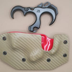 May include: A black archery release aid with a curved design, positioned above a tan holster with a red interior. The holster has a carbon fiber weave pattern and is secured with black rivets.