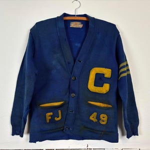 May include: A vintage navy blue cardigan sweater with a button-up front. The sweater features a large yellow "C" patch, yellow stripes on the sleeve, and yellow patches with "FJ" and "49". The sweater is hanging on a hanger.