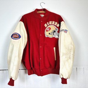 May include: A red varsity jacket with cream-colored sleeves, featuring a San Francisco 49ers helmet logo and the text NINERS on the chest. The right sleeve has an NFC patch, and the left sleeve displays an NFL logo. It has a snap-button front.