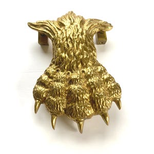 May include: A gold-colored metal door knocker shaped like a large animal paw with textured fur detailing on the upper section and five distinct claws extending from the base. The object features two mounting brackets visible at the top corners.