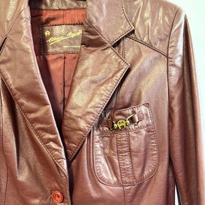 Pode incluir: Uma jaqueta de couro bord&ocirc; com um detalhe de fivela de metal dourado no bolso. A jaqueta tem um fecho de bot&atilde;o &uacute;nico e uma etiqueta que diz "Anne Klein" Feito na Coreia.