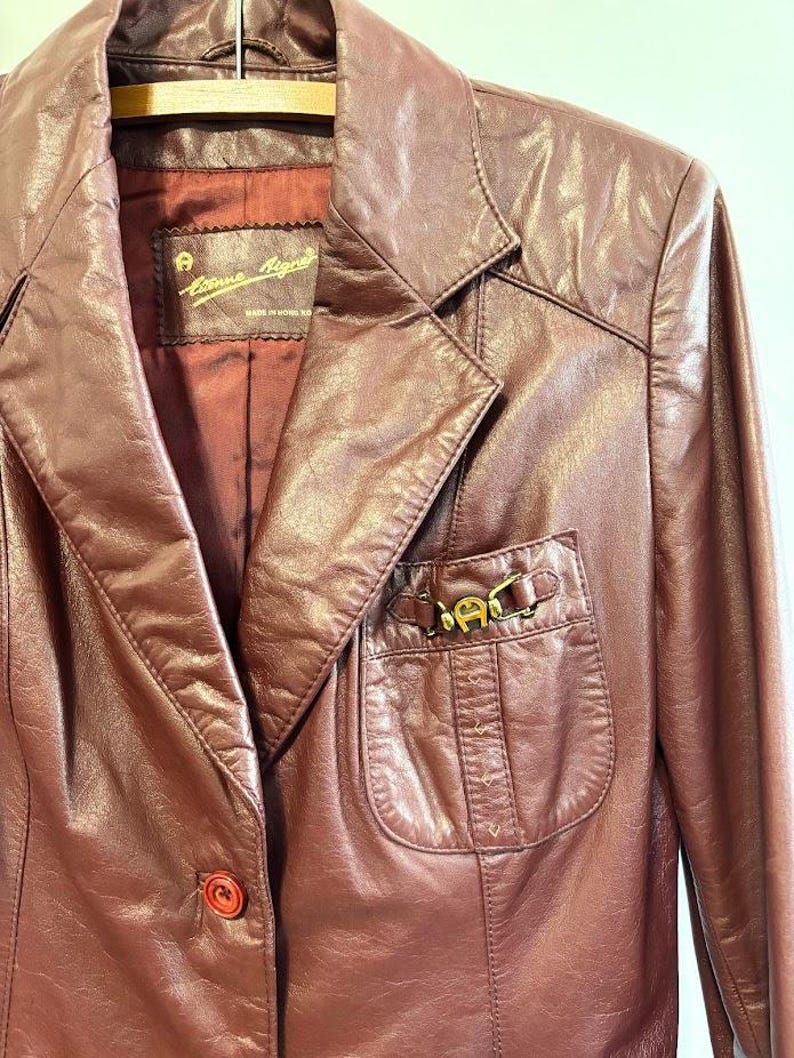 Pode incluir: Uma jaqueta de couro bord&ocirc; com um detalhe de fivela de metal dourado no bolso. A jaqueta tem um fecho de bot&atilde;o &uacute;nico e uma etiqueta que diz "Anne Klein" Feito na Coreia.