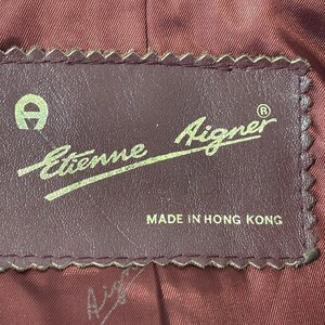Pode incluir: Uma etiqueta de couro marrom com costuras brancas e o texto "Etienne Aigner" em uma fonte cursiva. A etiqueta tamb&eacute;m inclui um logotipo "A" estilizado e o texto "Feito em Hong Kong".