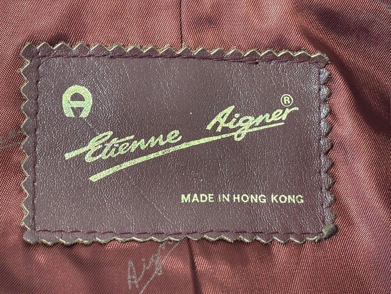 Pode incluir: Uma etiqueta de couro marrom com costuras brancas e o texto "Etienne Aigner" em uma fonte cursiva. A etiqueta tamb&eacute;m inclui um logotipo "A" estilizado e o texto "Feito em Hong Kong".
