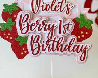 May include: A cake topper featuring red glitter strawberries with green glitter leaves and yellow dots. The text reads Violet's Berry 1st Birthday in red glitter script lettering layered over a light pink banner and white cardstock backing.