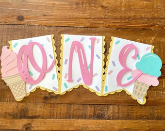 Könnte beinhalten: Ein dreiteiliges Papierbanner mit der Aufschrift ONE. Jeder weiße Wimpel ist mit pastellfarbenen Streuseln, einem gewellten gelben Rand und einem großen rosa Buchstaben versehen. Das Banner enthält Eiswaffel-Ausschnitte in Rosa und Türkis.