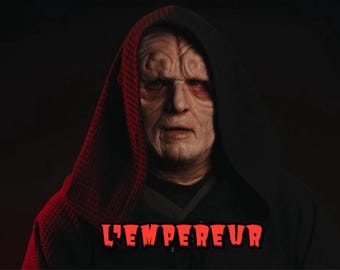 May include: A character with pale, wrinkled skin and deep-set eyes wearing a dark hooded robe with a textured red lining. The text L'EMPEREUR is displayed in bold red letters at the bottom center of the image against the dark clothing.