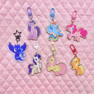 May include: A selection of My Little Pony keyrings. Each keyring showcases a different pony character, with a metal clasp for attachment. The keyrings are crafted from wood and acrylic, featuring bright colours and detailed designs. The background is a pink quilted fabric.