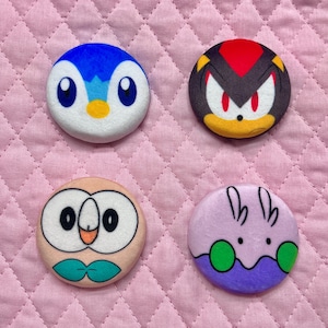 May include: Four round, colorful character buttons. One button features a blue penguin with a yellow beak. Another depicts a black and red character with a yellow muzzle. The other two buttons feature a light brown owl and a purple creature.