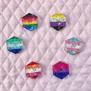 Può includere: Sei dadi poliedrici a venti facce, ciascuno con una diversa combinazione di colori, tra cui arcobaleno, viola e rosa. I dadi mostrano i numeri 2, 8, 14 e 20. I dadi sono disposti su una superficie trapuntata rosa.