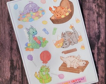 May include: A sheet of stickers featuring colorful cartoon dragons with candy themes. The stickers include a dragon eating candy corn, a dragon in a candy bar, a dragon with a lollipop, a dragon with a candy cane, and a dragon with a chocolate bar. The stickers are labeled "Halloween Candy Critters by Cat by Cider".