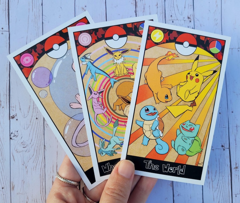 May include: Three illustrated Pokemon tarot cards with a white background. The cards are "The World", "Wheel of Fortune", and "The Moon". The cards feature various Pokemon characters, including Pikachu, Squirtle, and Eevee.