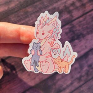 May include: A pink dragon with blue and white wings holding a black cat. A smaller orange cat sits to the right of the dragon. The sticker is on a wooden background.