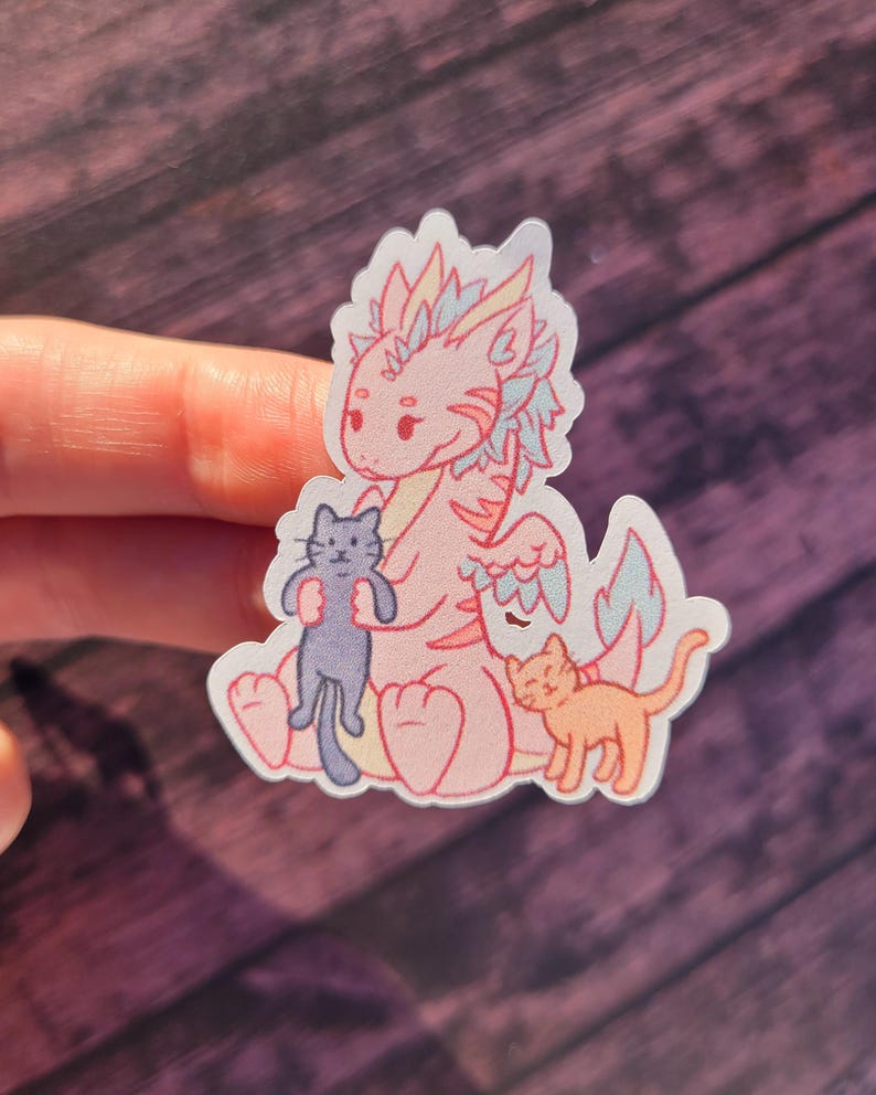 May include: A pink dragon with blue and white wings holding a black cat. A smaller orange cat sits to the right of the dragon. The sticker is on a wooden background.