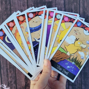 May include: A set of tarot cards featuring illustrations of various Pokemon characters. The cards are spread out in a fan shape, with the card labeled "The Fool" visible in the foreground. The Fool card features a yellow Pokemon with a blue water droplet on its head.