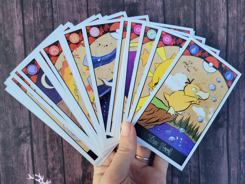 May include: A set of tarot cards featuring illustrations of various Pokemon characters. The cards are spread out in a fan shape, with the card labeled "The Fool" visible in the foreground. The Fool card features a yellow Pokemon with a blue water droplet on its head.