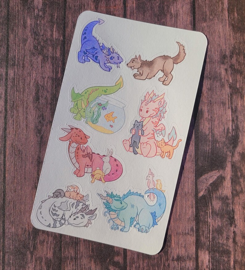 May include: A sheet of stickers featuring various cute cartoon dragons, cats, and a dog. The dragons are pink, blue, and green, and the animals are holding or playing with other animals. The stickers are on a white background.