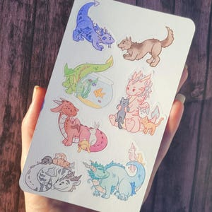 May include: A sheet of stickers featuring various cartoon dragons, a wolf, and a fishbowl with fish. The dragons are in different colors, including blue, green, pink, and red. The stickers are on a white background.