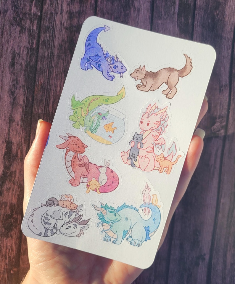 May include: A sheet of stickers featuring various cartoon dragons, a wolf, and a fishbowl with fish. The dragons are in different colors, including blue, green, pink, and red. The stickers are on a white background.