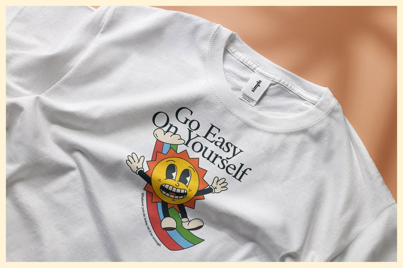 Puede incluir: Camiseta blanca con un gr&aacute;fico de un sol sonriente con un arco&iacute;ris y el texto "Go Easy On Yourself".