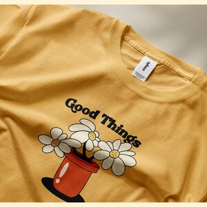 Pu&ograve; includere: Una maglietta color senape con una grafica di un vaso rosso con margherite bianche e il testo "Good Things".