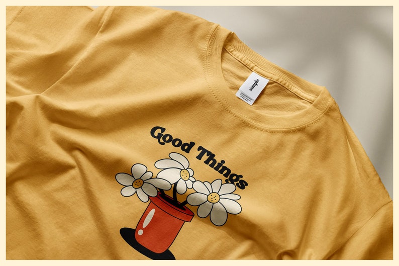 Pu&ograve; includere: Una maglietta color senape con una grafica di un vaso rosso con margherite bianche e il testo "Good Things".