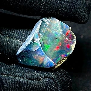 Puede incluir: Una piedra preciosa de ópalo en bruto con un color base oscuro, que muestra vibrantes destellos de verde, rojo y azul. La piedra tiene una superficie pulida e iridiscente y un borde natural y en bruto. El ópalo se sostiene sobre un fondo negro.
