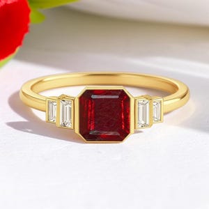 May include: A gold ring featuring a central emerald-cut deep red gemstone flanked by two small baguette-cut clear stones on each side. The band is smooth and polished, showcasing a classic design with a vibrant contrast between the red and gold.