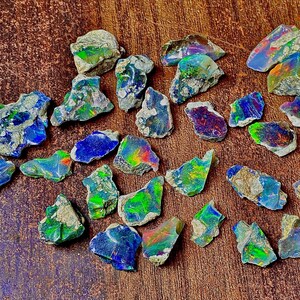 Puede incluir: Una colección de piedras preciosas de ópalo en bruto, que muestra una vibrante gama de colores, incluyendo azules, verdes y destellos de tonos arcoíris. Los ópalos están tallados en bruto, mostrando su estado natural sin pulir. Las piedras preciosas están dispersas sobre una superficie de madera.