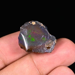 Könnte beinhalten: Ein rauer, ungeschliffener schwarzer Opal-Edelstein mit lebendigen grünen, roten und blauen Blitzen. Der Opal wird in einer Hand gehalten, wodurch seine natürliche, unregelmäßige Form und sein einzigartiges Farbspiel hervorgehoben werden. Der Stein hat eine dunkle, fast schwarze Grundfarbe.