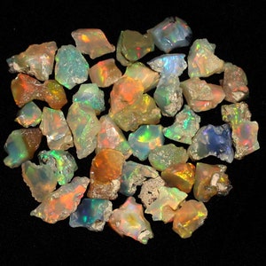 Puede incluir: Una colección de piedras preciosas de ópalo en bruto e iridiscente en varios tonos de verde, azul, naranja y rojo. Los ópalos tienen una forma irregular y un acabado natural sin pulir.