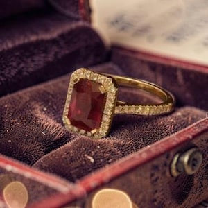 Puede incluir: Un anillo de oro con una gran gema roja rectangular, rodeada de diamantes más pequeños. El anillo se exhibe en un joyero de terciopelo granate con tapa abisagrada.