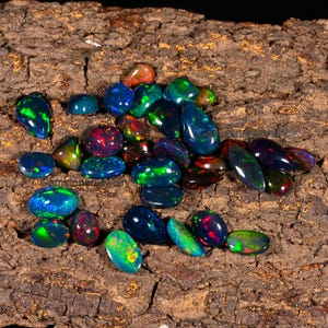 May include: A collection of black opal gemstones in various shapes and sizes, with iridescent colors of green, blue, red, and purple. The gemstones are displayed on a piece of rough-hewn wood.