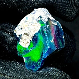 Puede incluir: Una piedra preciosa de ópalo en bruto con colores vibrantes. La piedra muestra una mezcla de verde brillante, azul profundo y toques de rojo y púrpura. La parte superior del ópalo tiene una superficie rugosa, blanca y texturizada, que contrasta con las facetas pulidas y coloridas.