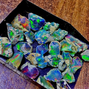 以下が含まれることがあります： 黒いトレイに入った、原石のオパール。オパールは青、緑、赤、紫などの鮮やかな色を示しています。宝石は自然で未研磨の外観で、滑らかで質感のある表面が混在しています。「thegemstonehub19」の文字が見えます。