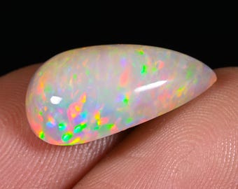 Könnte beinhalten: Ein polierter, tropfenförmiger Opal-Edelstein. Der Opal zeigt eine milchig-weiße Grundfarbe mit lebendigen Blitzen in Grün, Gelb, Orange und Rosa. Der Edelstein wird vor einem dunklen Hintergrund gehalten, was sein schillerndes Farbspiel hervorhebt.